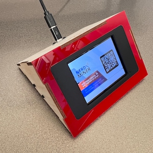 May include: An angled electronic device with a red frame and a screen displaying a QR code and the text "NERD MINER". The device has a wooden base and a USB cable plugged in. The screen also displays "WAITING CONFIG... USE THIS WIFI TO SETUP".