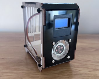 May include: A small, clear acrylic case with a black front panel. The front panel has a small LCD screen and a fan with a black grill. The case is sitting on a wooden surface.