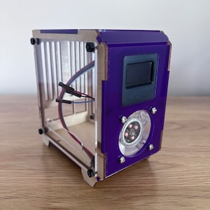 May include: A small, clear acrylic case with a purple front panel. The case has a small fan with a black screen on the front. The case is sitting on a wooden surface.
