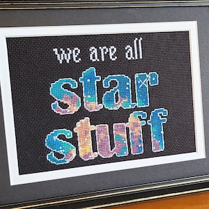 May include: A framed cross-stitch with the text "we are all star stuff" in colorful embroidery. The frame is black with a gold trim. A book titled "Cosmos Carl Sagan" is visible in the background.