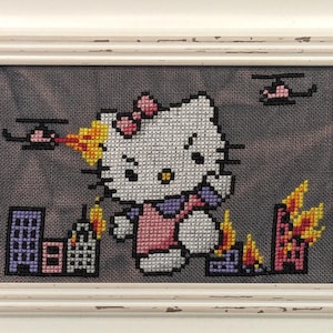 Puede incluir: Arte de punto de cruz de un personaje de gato de dibujos animados con un vestido rosa, destruyendo una ciudad. La obra enmarcada presenta un marco blanco desgastado. Los edificios están en llamas y hay helicópteros en el cielo.
