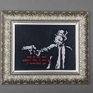 May include: A framed cross-stitch artwork featuring a black and white image of a person holding a gun. The text "Pass go, one more goddamn time. I dare you. I double-dare you." is written in red below the image.