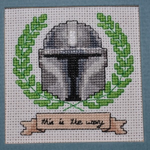 May include: Cross-stitch embroidery of a Mandalorian helmet with a green laurel wreath and the text "This is the way".