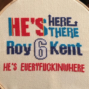 Peut inclure: Un cercle de broderie au point de croix avec le texte "HE'S HERE THERE Roy 6 Kent HE'S EVERYFUCKINGWHERE" en rouge, bleu et blanc. La broderie est sur un tissu crème, et le cercle est en bois.