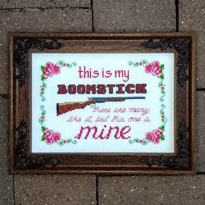 May include: A framed cross-stitch design with a brown wooden frame. The cross-stitch features a pink and green floral border with the text "this is my BOOMSTICK there are many like it, but this one is mine".