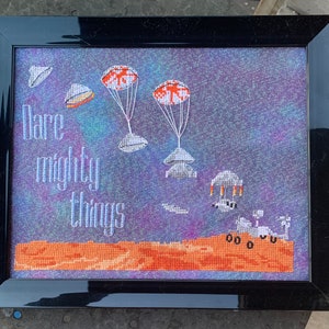 May include: A framed cross-stitch design with a purple and blue background. The design features a rocket ship landing on a red planet with the text "Dare mighty things" in white.