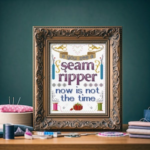 May include: A framed cross-stitch design with a gold frame. The cross-stitch features a colourful design of sewing tools and the text "If you see a seam ripper now is not the time."