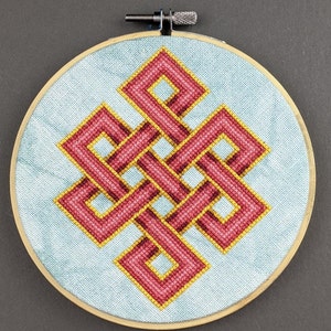Pode incluir: Um aro de bordado circular com um fundo de tecido azul e um desenho de nó infinito bordado em vermelho e dourado.