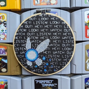 May include: A black and white embroidered hoop with a pixelated image of a blue and white character from the video game "Star Fox 64". The text "HEY! LISTEN! LOOK! WATCH OUT!" is embroidered around the character.