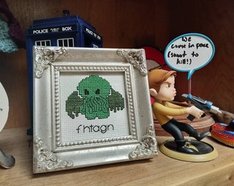 May include: A silver framed cross-stitch with a green Cthulhu monster and the text "fhtagn". A Star Trek figurine with a speech bubble that says "We come in peace (shoot to kill!)" is in the background.