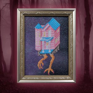 Puede incluir: Arte de punto de cruz de una casa rosa con tejado azul y patas de pollo, sobre un fondo morado oscuro. La obra está enmarcada en un marco plateado ornamentado. La casa tiene varias ventanas y un porche blanco.