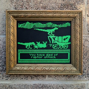 Pode incluir: Uma obra de arte de ponto cruz enquadrada que retrata um dinossauro pixelado verde atacando um jipe. O texto "You have died of raptor attack" é exibido em uma caixa verde abaixo da imagem.