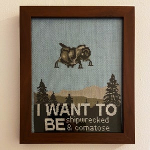 May include: A framed cross-stitch with a blue background and a small spaceship in the center. The text below reads "I want to be shipwrecked & comatose".