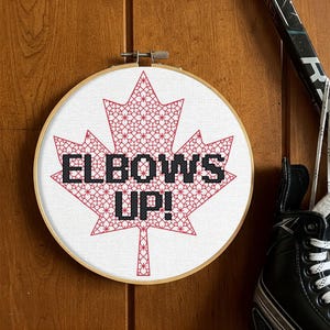 May include: Embroidered red maple leaf with the text "Elbows Up!" on a white background in a wooden embroidery hoop.