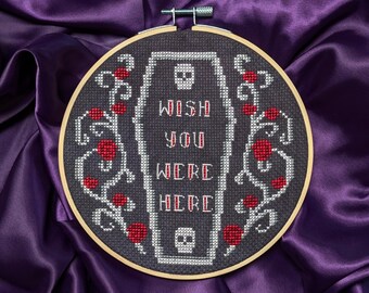 May include: A cross-stitch embroidery in a wooden hoop. The design features a coffin with the words "WISH YOU WERE HERE" in red and white. Red roses and vines surround the coffin, with a skull at the top and bottom. The background is dark gray.