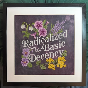 Könnte beinhalten: Gerahmtes Kreuzstich-Kunstwerk mit dem Text "Radicalized by Basic Decency", umgeben von floralen Stickereien. Die Blumen umfassen lila Petunien, Lavendel, gelbe Blüten und weiße Schneeglöckchen. Das Kunstwerk ist in einem schwarzen Rahmen mit einem weißen Passepartout gerahmt.