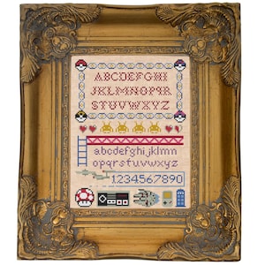 May include: A framed cross-stitch design featuring a variety of retro video game and pop culture icons. The design includes the alphabet, numbers, and images of characters from Pac-Man, Super Mario Bros., Star Wars, and Doctor Who.