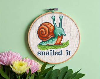 May include: A cross-stitch embroidery hoop with a green snail with a brown shell on a green leaf. The text "snailed it" is below the snail.