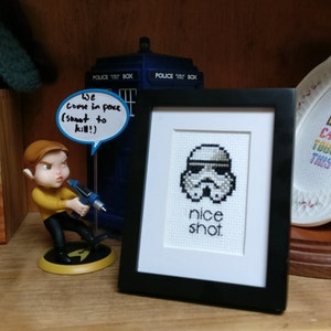 May include: A framed cross-stitch featuring a Stormtrooper helmet with the words "nice shot." A figurine of a character holding a weapon is to the left. A blue police box is in the background.