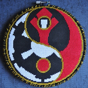 May include: A round, black and white embroidery hoop with a red and gold design. The design features a yin yang symbol with a red circle in the center of the black half and a red arrow pointing up in the center of the white half.