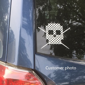 May include: A white vinyl sticker of a cross-stitch skull with two needles crossed behind it. The sticker is on the back window of a blue car. The text "Customer photo" is visible in the lower right corner of the image.