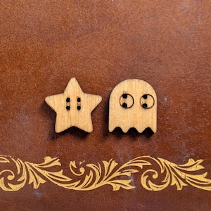 Puede incluir: Dos botones de madera: una estrella con dos ojales y un fantasma con dos agujeros para los ojos. Los botones son de color marrón claro y están colocados sobre un fondo marrón con un borde decorativo dorado.