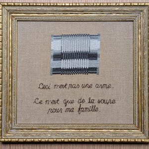 May include: A framed cross-stitch with a black and white geometric design. The text below the design reads "Ceci n'est pas une arme. Ce n'est que de la soure pour ma famille."
