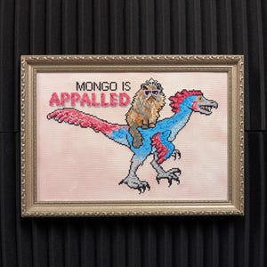 Könnte beinhalten: Kreuzstichkunst in einem goldenen Rahmen. Das Kunstwerk zeigt eine Katze mit Sonnenbrille und Krone, die auf einem blau-roten Dinosaurier reitet. Der Text "MONGO IS APPALLED" steht über der Katze und dem Dinosaurier.