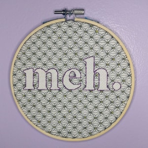 May include: A round wooden embroidery hoop with a white fabric background and a black and white geometric pattern. The word "meh." is embroidered in white with a purple outline.