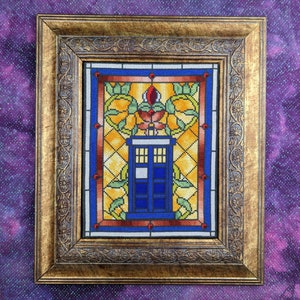 May include: Cross-stitch artwork of a blue police box, framed in a gold-coloured, ornate wooden frame. The artwork features a stained-glass style design with yellow, orange, red, and green accents. The background is a purple fabric.