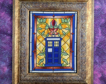 May include: Cross-stitch artwork of a blue police box, framed in a gold-colored, ornate wooden frame. The artwork features a stained-glass style design with yellow, orange, red, and green accents. The background is a purple fabric.