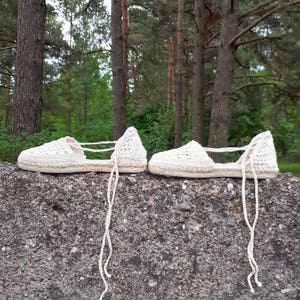 May include: A pair of white crocheted sandals with a woven jute sole. The sandals have a lace-up design and are tied at the ankle.