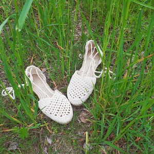 May include: Two white crocheted sandals with a delicate pattern, lying on a bed of green grass.