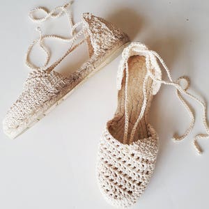 May include: A pair of cream-coloured crocheted espadrille sandals with lace-up ties. The sandals have a woven texture and a flat sole.
