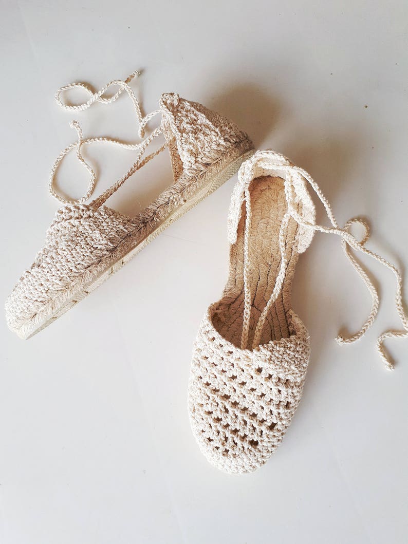 May include: A pair of cream-coloured crocheted espadrille sandals with lace-up ties. The sandals have a woven texture and a flat sole.