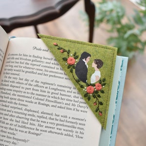 May include: A green felt bookmark with embroidered flowers and a picture of a couple. The bookmark is folded into a triangle shape and has the text "Pride and" on the front.