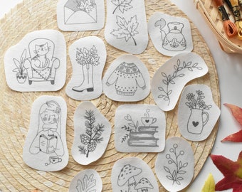 Pode incluir: Um conjunto de 14 desenhos de transferência termoadesiva com ilustrações de temática outonal. Os desenhos incluem um gato numa cadeira, botas, um suéter, um bule, uma caneca de chocolate quente, um ramo de flores, um cogumelo, uma folha, um ramo de pinho, uma pilha de livros e um envelope. As transferências estão sobre um fundo branco e estão prontas para serem passadas a ferro em tecido.