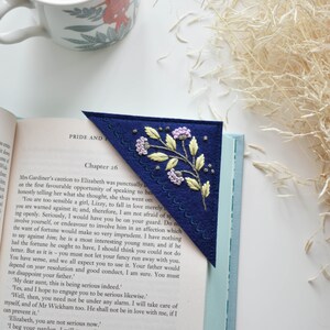 May include: Navy blue felt bookmark with embroidered floral design in yellow, light green, and purple. The bookmark is placed in a book with the title "Pride and Prejudice" visible. A cup of coffee sits nearby.