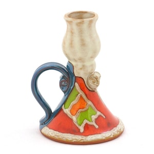 Puede incluir: Un portavelas de cerámica con un asa azul y un diseño geométrico colorido. El portavelas está pintado en tonos de rojo, naranja, verde y blanco.