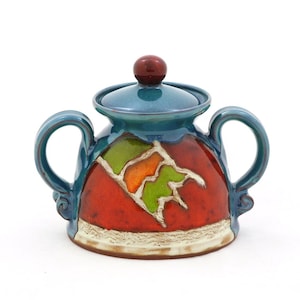 Może przedstawiać: Ceramiczna cukiernica z pokrywką i dwoma uchwytami. Miska jest turkusowa z czerwonym i zielonym wzorem. Pokrywka ma mały, okrągły, ciemnoczerwony guz. Podstawa miski ma teksturowany, kremowy pasek.