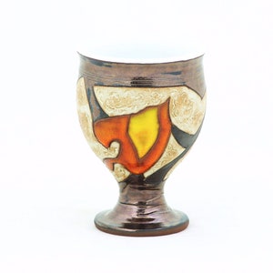 May include: A ceramic goblet with a brown, white, orange, and yellow geometric design. The goblet has a wide base and a rounded bowl.