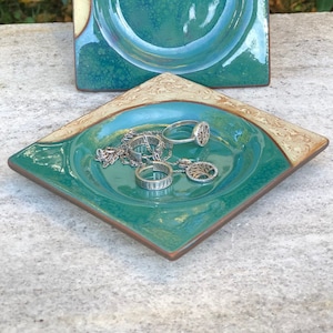 Puede incluir: Dos platos cuadrados de cerámica con un esmalte turquesa y un borde elevado. El plato más pequeño tiene un anillo, un collar y un colgante dentro.