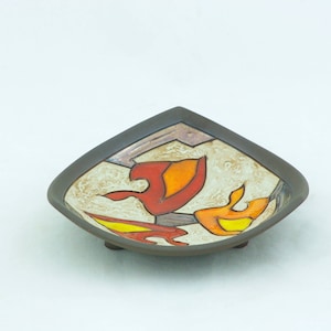 以下が含まれることがあります： 白地に赤、オレンジ、黄色のスタイリッシュな花柄が施された、3本の足が付いた正方形の陶器製の皿。皿は濃い茶色の縁取りがされている。