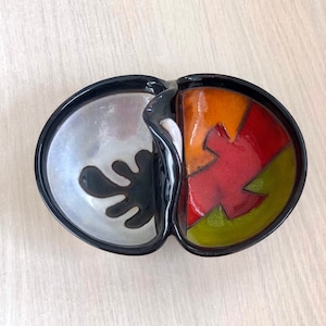 Puede incluir: Un plato dividido de cerámica negra con dos secciones. Una sección es blanca con un diseño de salpicaduras negras. La otra sección es multicolor con rojo, naranja, amarillo y verde.