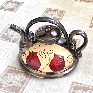 May include: A ceramic teapot with a brown glaze and red floral designs. The teapot has a round body and a curved spout. The lid is also ceramic and has a brown glaze.