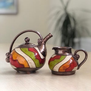 以下が含まれることがあります： 赤、オレンジ、緑、白の色合いでカラフルな幾何学模様が施された、セラミック製のティーポットとクリーマーのセット。ティーポットは湾曲した注ぎ口とハンドルが付いており、クリーマーは小さなハンドルが付いています。