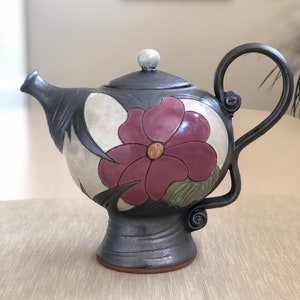 May include: A black ceramic teapot with a floral design. The teapot has a round body with a single handle and a lid with a knob. The floral design features a large pink flower with a yellow centre and green leaves.