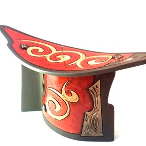 Puede incluir: Una escultura de cerámica roja y dorada con una forma curva y diseños intrincados. La escultura tiene una base negra y un alfiler dorado en el centro.