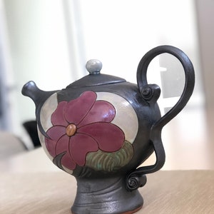 May include: A black ceramic teapot with a floral design. The teapot has a round body with a single handle and a spout. The floral design features a large pink flower with green leaves.