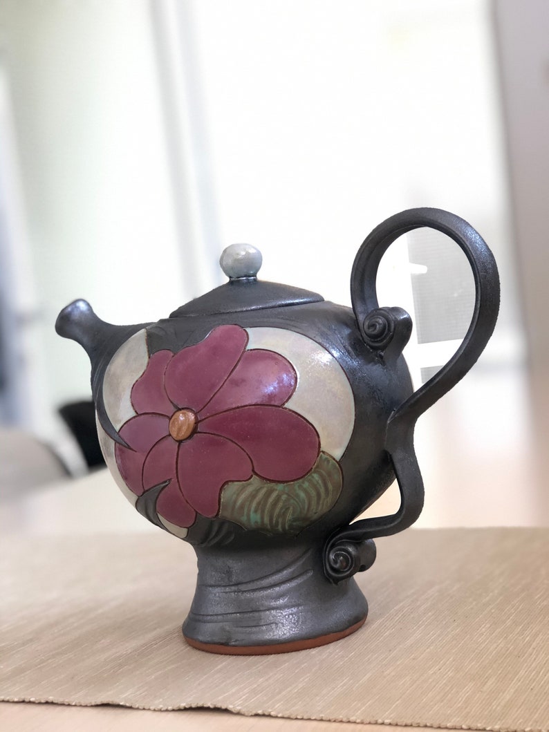 May include: A black ceramic teapot with a floral design. The teapot has a round body with a single handle and a spout. The floral design features a large pink flower with green leaves.